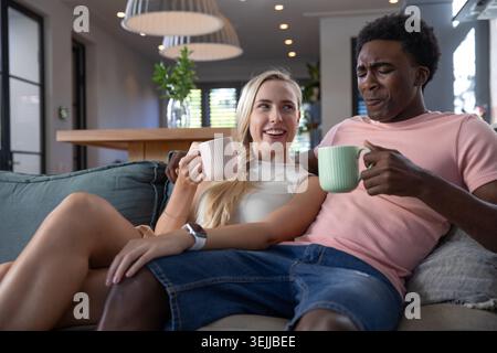 Couple diversifié sirotant des tasses pastel tout en étant assis sur un canapé gris par une table à manger en bois. Confort, intimité, détente, conte Banque D'Images