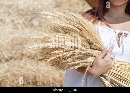 Jeune femme assise sur la botte de foin dans le champ récolté tenant le bouquet de blé Banque D'Images