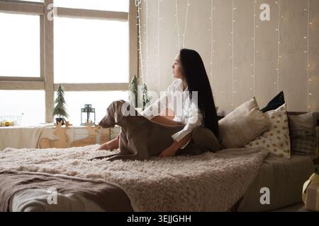 Heureuse jeune femme en pyjama avec chien weimaraner attendent le nouvel an à la maison, souriant et regardant la caméra. Banque D'Images