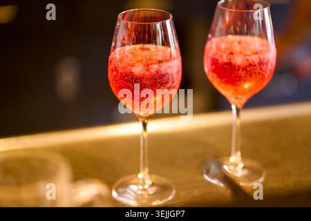 Deux verres de cocktail rouge pétillant avec de la glace, probablement Kir Royale ou Aperol Spritz, debout sur un comptoir de bar sous un éclairage chaleureux. Banque D'Images