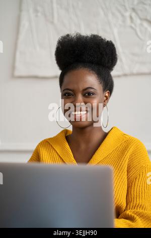 Femme millénaire afro-américaine joyeuse avec coiffure afro dans le cardigan jaune à distance travaillant sur ordinateur portable, regardant la caméra, h. Banque D'Images