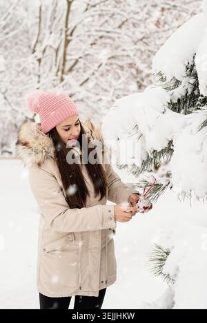 Belle femme dans des vêtements chauds d'hiver décorant l'arbre de Noël dans un parc en journée enneigée Banque D'Images