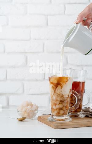 Main versant le lait dans le verre avec du thé à bulles glacé, fond de mur de briques blanches Banque D'Images