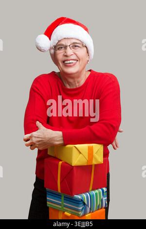 Heureuse dame senior dans le chapeau rouge Santa avec des cadeaux de Noël regardant la caméra et souriant posant sur un mur. Noël. Banque D'Images