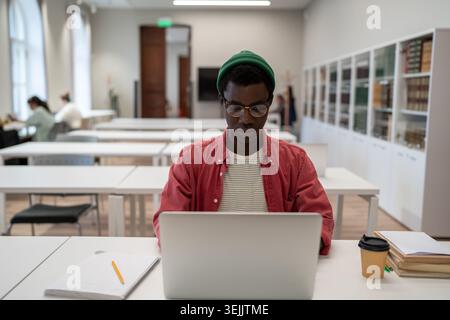Un homme afro-américain spécialisé dans les lunettes étudie en ligne sur un ordinateur portable tout en étant assis à la bibliothèque de l'université Banque D'Images