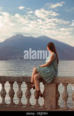 Femme assise sur une balustrade en pierre regardant le lac calme et les montagnes lointaines Banque D'Images