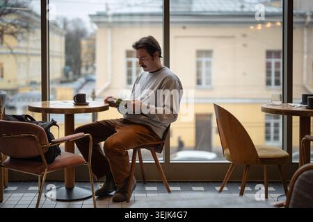Homme amoureux du livre entièrement engagé dans la lecture du livre populaire tout en étant assis dans un café confortable avec une tasse de café. Banque D'Images
