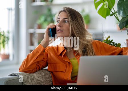 Femme d'âge moyen étourdie recevant des nouvelles choquantes soudaines via smartphone alors qu'elle était assise à l'ordinateur portable Banque D'Images