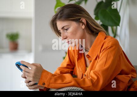 Vue latérale d'une femme d'âge moyen défilant intensément à travers le fil d'actualité sur le téléphone assis sur le canapé Banque D'Images