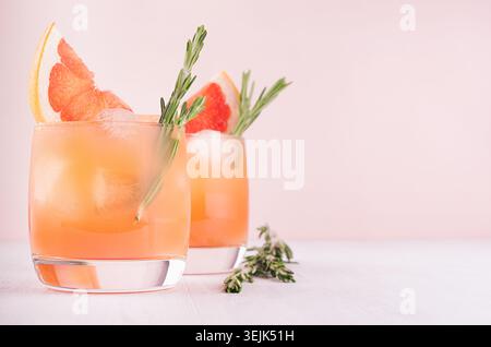 Cocktail détox froid de jus de pamplemousse avec glace, romarin, tranches d'agrumes sur fond rose clair et blanc doux. Banque D'Images
