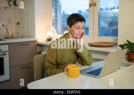 Heureuse femme d'affaires senior réfléchie regardant l'écran d'ordinateur portable avec une expression souriante positive. Banque D'Images