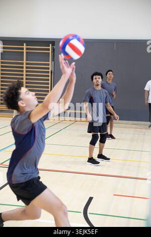 Équipe masculine de volleyball adolescente diversifiée atteignant et plaçant le volleyball tricolore sur le sol de la salle de gym de l'école. Cour, athlètes, coopération, Banque D'Images