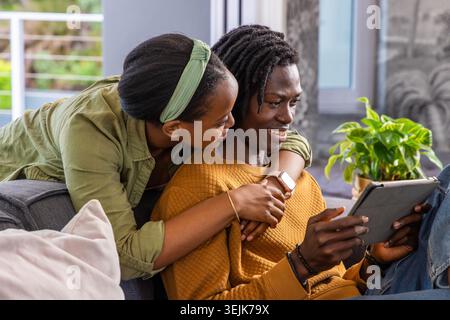 Couple afro-américain assis sur un canapé gris, homme tenant une tablette, femme penchée et embrassant. Appareil, plante, fenêtre, Balc Banque D'Images