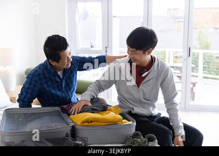 Père asiatique et fils adultes assis sur le lit emballant une valise ouverte avec un pull jaune et des écouteurs Banque D'Images