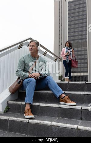 Navetteurs assis sur les marches près de la tour de bureaux, homme portant un casque utilisant une tablette, femme tenant une tasse. Urbain, escalier, architecture, Banque D'Images