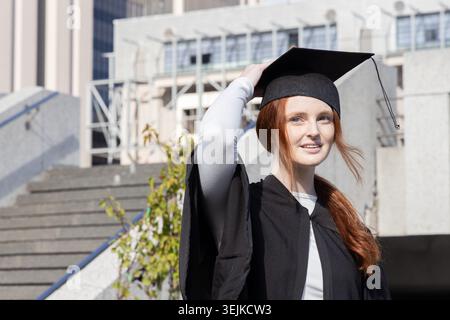 Femme diplômée debout sur des marches en béton sur le campus dans la casquette et la robe tenant mortarboard, espace de copie. Université, escalier, rega Banque D'Images