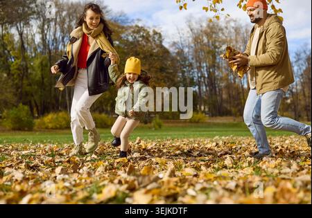 Parents souriants et fille courant à travers les feuilles d'automne dans le parc ensoleillé de la ville Banque D'Images