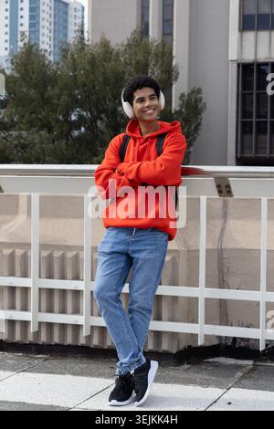 Afro-américain adolescent masculin appuyé sur le parapet sur le toit, souriant en sweat à capuche rouge et sac à dos noir. Urban, écouteurs, sac, snea Banque D'Images