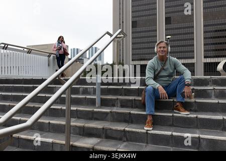 Homme senior assis sur les marches de la plaza portant des écouteurs et tenant le café pendant que la femme asiatique appelle. Piétons, urbains, navetteurs, Banque D'Images