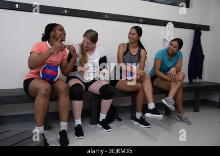Diverses coéquipières féminines de volley-ball assis sur un banc en bois dans le vestiaire contenant des balles de volley-ball. Équipe, ballon, siège, camaraderie, a Banque D'Images