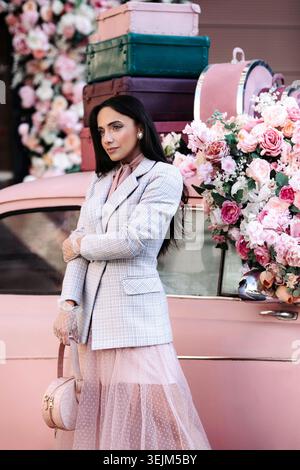 Modèle de dame de mode en robe de veste rose, maquillage et cheveux noirs sur voiture rétro décorée de roses Banque D'Images
