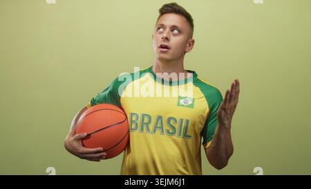 Jeune homme blond tient le basket-ball sous le bras et fait des gestes de la main contre la poitrine tout en portant un maillot brésilien en studio ; fierté de l'équipe. Banque D'Images