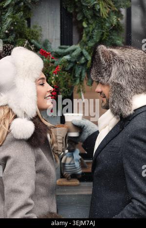 Un couple d'amateurs de mode se tenant sur la rue d'hiver par temps enneigé, souriant, buvant des boissons chaudes. Banque D'Images