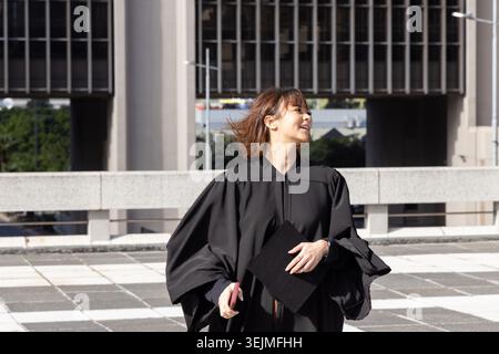 Diplômée asiatique debout sur la place carrelée portant une casquette de maintien de robe noire et un smartphone rose. Rooftop, extérieur, urbain, Terra Banque D'Images