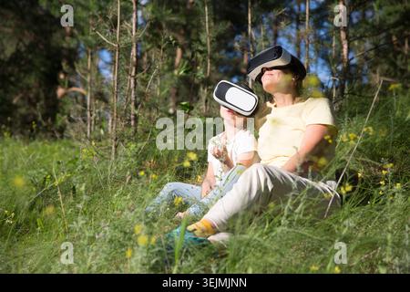 Maman et fille d'enfant jouant au jeu dans des lunettes de réalité virtuelle et détendu assis sur l'herbe dans la forêt à l'extérieur. copier Banque D'Images