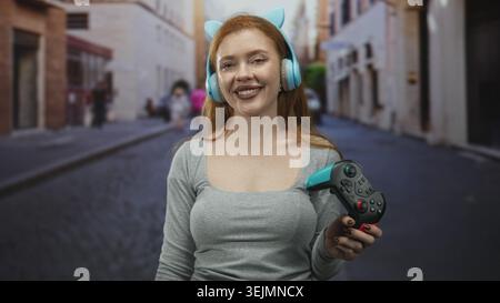 Femme tenant une manette de jeu et souriant les yeux fermés tout en portant des écouteurs turquoise dans une rue ; contentement de jeu décontracté. Banque D'Images