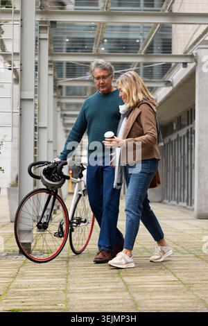 Homme senior et femme marchant sous la verrière tenant le vélo de route avec casque tout en sirotant du café. Urbain, passerelle, architecture Banque D'Images