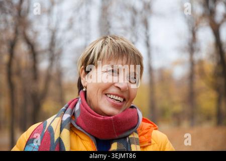 Joyeux rire femme senior dans une écharpe et veste regarde dans la caméra marche à travers le parc d'automne. bannière Banque D'Images