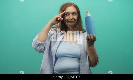 Femme senior tient une bouteille de savon bleue tout en pointant l'index vers la tempe dans le studio ; démonstration de produit réfléchie. Banque D'Images