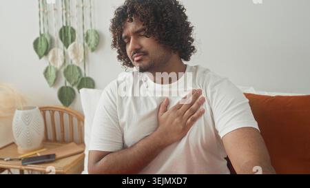Jeune homme indien serrant la poitrine et appuyant main contre poitrine sur le lit dans le studio avec table de chevet et smartphone, suggérant heartattack ; douleur. Banque D'Images