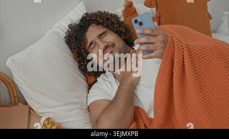 Jeune homme indien couché dans le lit sous une couverture orange tient smartphone, touche la gorge avec la main et gagne à côté de la bouteille de pilule dans le studio ; problème de santé. Banque D'Images