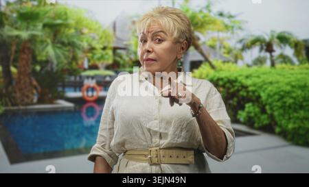 Femme hispanique senior pointant le doigt avec une main visible et des anneaux à côté de la piscine du complexe portant une chemise en lin et une ceinture ; avertissement de poupe. Banque D'Images