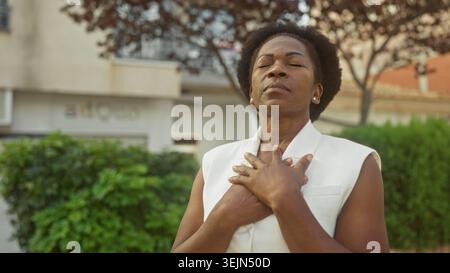 Femme avec les mains pressées sur la poitrine et les yeux fermés debout devant une façade de bâtiment à côté de haies dans la rue ; sérénité. Banque D'Images