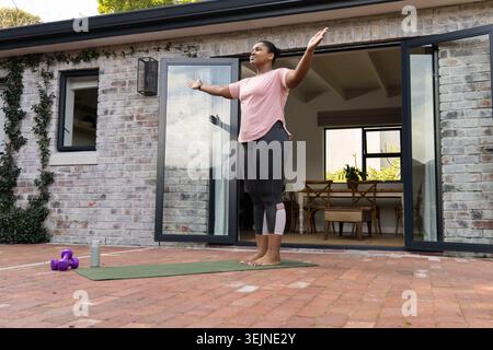 Femme afro-américaine s'étirant sur un tapis de yoga, patio en briques rouges à côté de portes coulissantes, en tee-shirt rose. Pieds nus, terrasse, extérieur, fi Banque D'Images