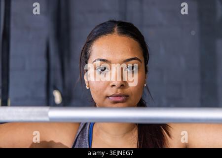 Femme debout derrière une barre horizontale au gymnase, portant un débardeur gris et une sangle bleue. Force, concentration, athlétisme, forme physique, tr Banque D'Images