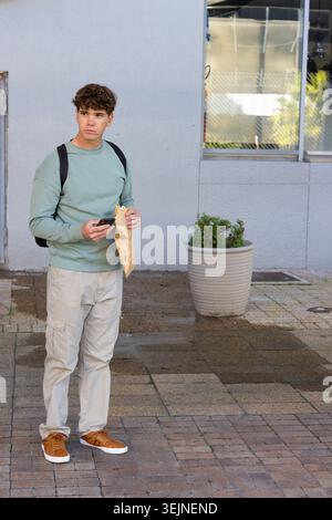 Adolescent homme debout sur la plaza tenant un smartphone et un sac en papier, portant un dessus de menthe et un sac à dos. Urbain, trottoir, minimaliste, gagnant Banque D'Images