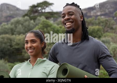 Couple diversifié debout sur le sentier tenant des tapis de yoga verts tout en souriant dans des vêtements athlétiques. Extérieur, nature, fitness, bien-être, PA Banque D'Images