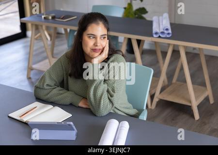 Femme assise en studio portant un pull vert, regardant pensive et utilisant un cahier, des plans roulés. Concepteur, dessin, plans, Banque D'Images