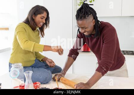 Couple diversifié faisant de la pâte sur l'îlot de cuisine, femme saupoudrant de la farine, homme utilisant un rouleau à pâtisserie en bois. Homestyle, comptoir, mode Banque D'Images