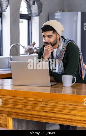 Bonnet d'homme indien et écharpe assis à l'îlot de cuisine travaillant sur un ordinateur portable argenté avec une tasse blanche. Espace de travail, bureau à domicile, minimal, c Banque D'Images