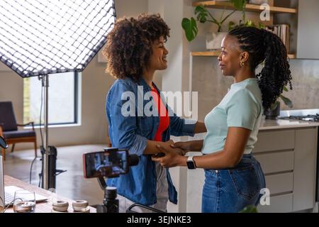 Femmes afro-américaines se tenant la main, parlant dans un studio de cuisine avec softbox et trépied de smartphone. Duo, créateurs, collaboratio Banque D'Images