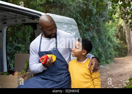 Homme afro-américain portant un tablier, garçon assis sur le pare-chocs de van au chemin, tenant des poivrons près de la caisse. Extérieur, marché, famille, ferme Banque D'Images
