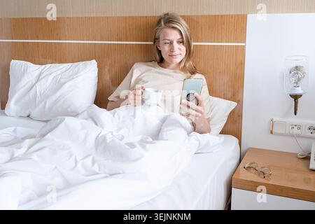 Femme utilisant un smartphone couché dans le lit et buvant du café Banque D'Images