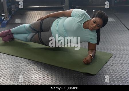 Femme afro-américaine exécutant planche latérale sur tapis vert au gymnase portant smartwatch, chaussures marron. Athlète, fitness, entraînement, je Banque D'Images