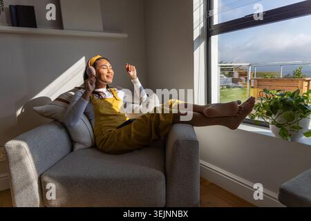 Femme asiatique s'inclinant dans un fauteuil à la maison portant une combinaison jaune écoutant avec un casque, téléphone sur les genoux. Sunlight, écouteurs, tablette, fenêtre, plante, confortable, Banque D'Images
