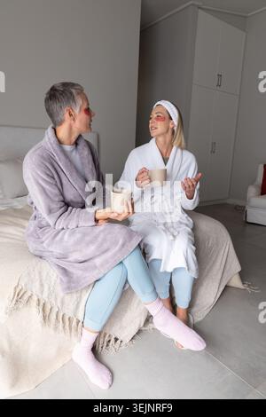 Amies âgées et d'âge moyen assises sur le lit portant des cache-yeux et des robes tenant des tasses. Paire, amitié, confortable, ambiant, Banque D'Images
